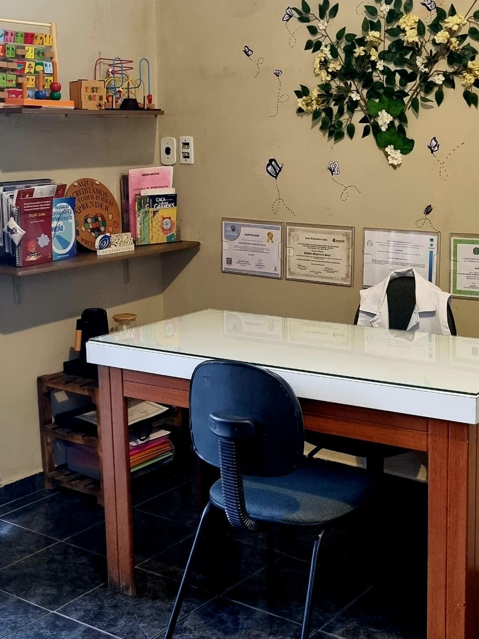 Sala de atendimento com mesa, cadeiras, prateleiras de jogos e livros pedagógicos e os certificados na parede