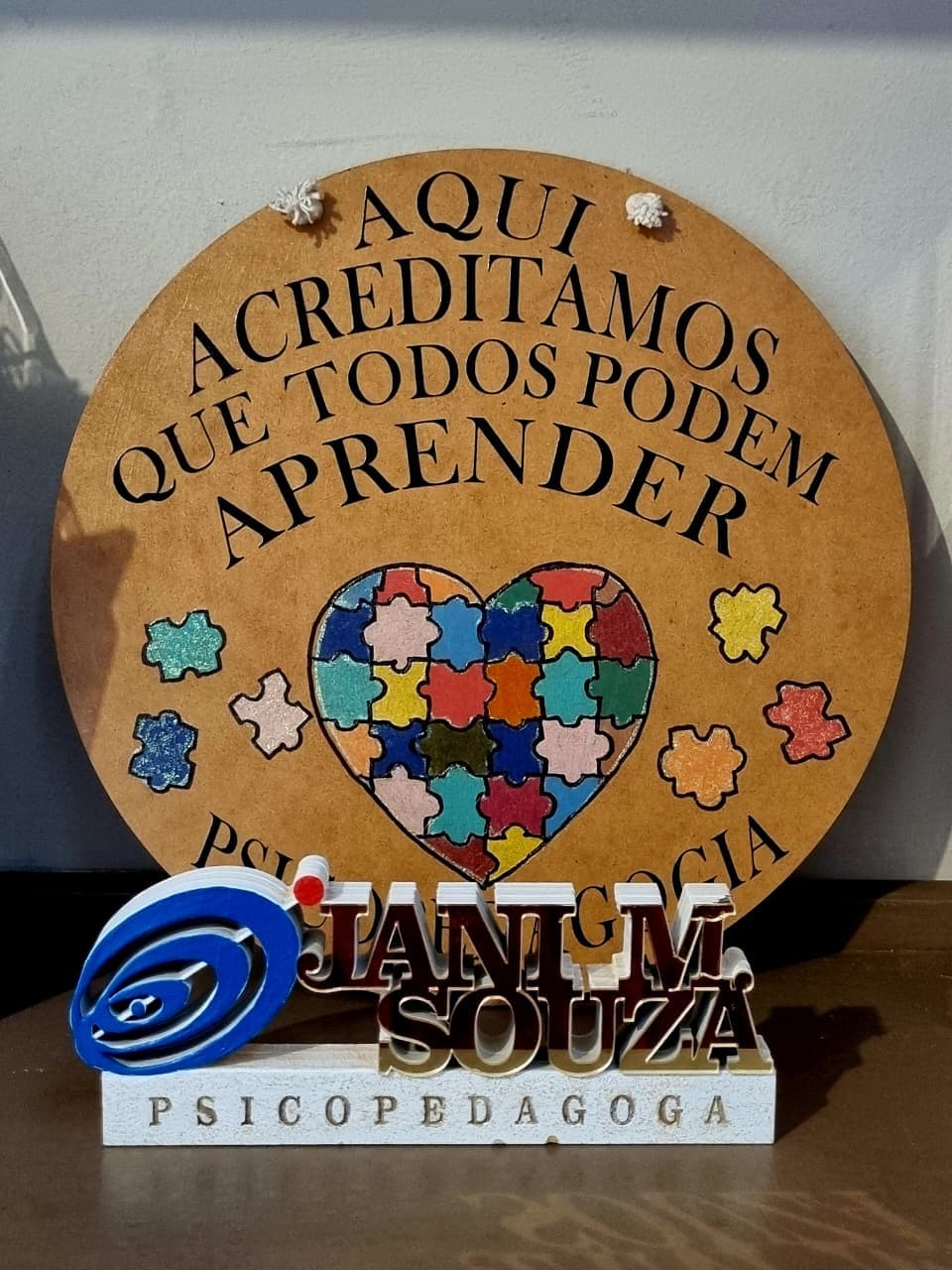 Placa de MDF na parede do consultório com a frase “Aqui acreditamos que todos podem aprender”, e a plaquinha de mesa “Jani M. Souza, Psicopedagoga”